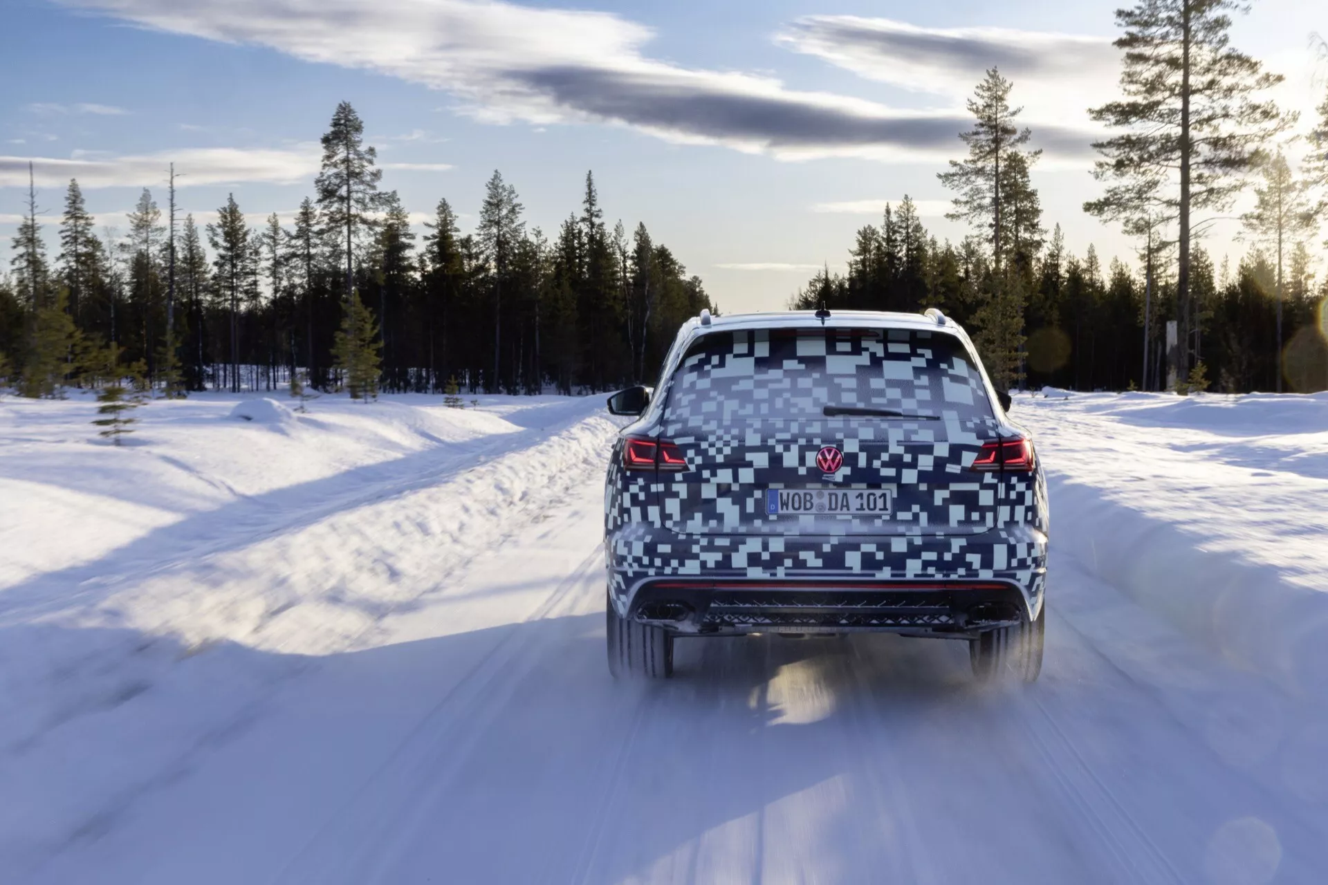 2023 Volkswagen Touareg (III-Facelift) rear view driving on snow-covered road in winter forest, showing glowing taillights, clear rear windshield, dual exhausts, and roof spoiler.