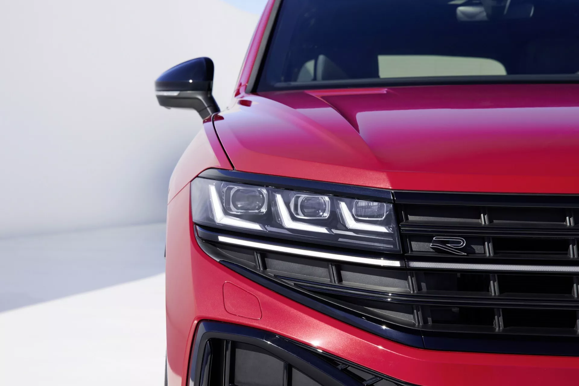 2023 VW Touareg III-Facelift with glossy metallic red paint featuring close-up view of the front left quarter, showing the LED headlight assembly, part of the horizontal slat grille, detailed view of the front bumper, and aerodynamic lines on the hood, against a blurred background emphasizing the car’s modern design and technological features.